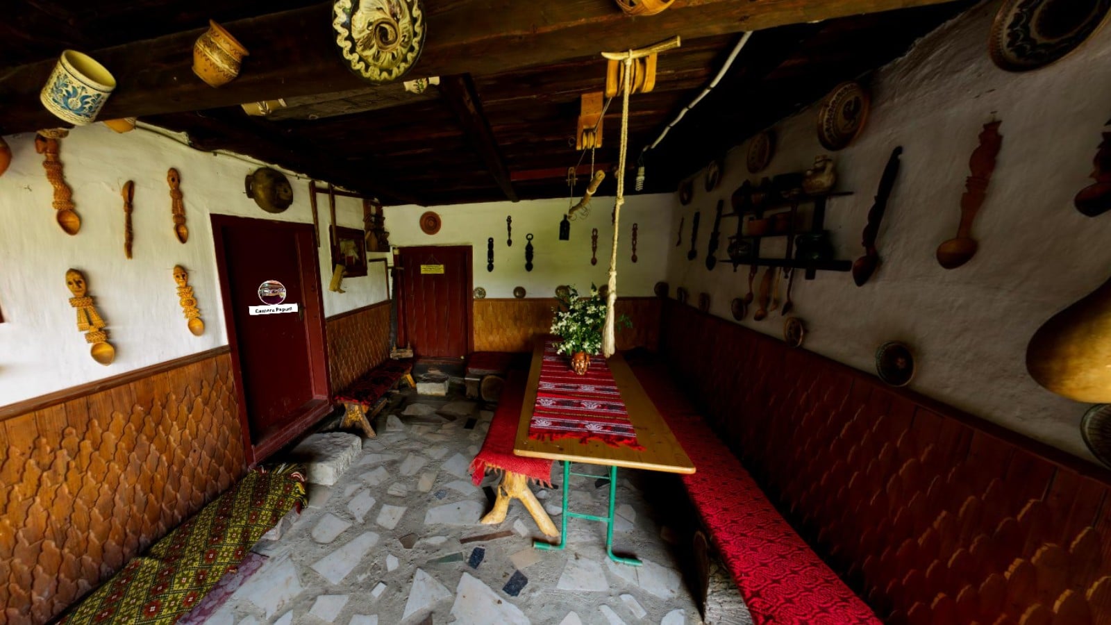 Interior traditional al unei case țărănești românești, cu masă lungă, bănci cu textile roșii și ceramică expusă pe pereți.
